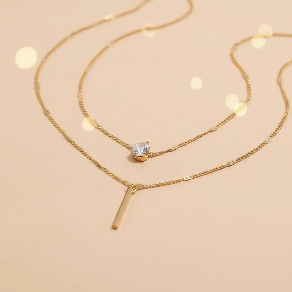 Close-up of gold Star Layered Necklace chains showing sparkling round crystal charm and sleek vertical bar pendant on peach backdrop | Oria Jewel

