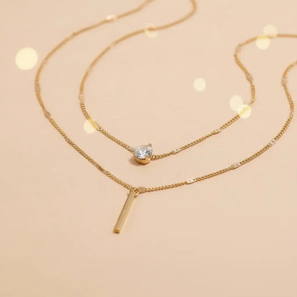 Close-up of gold Star Layered Necklace chains showing sparkling round crystal charm and sleek vertical bar pendant on peach backdrop | Oria Jewel

