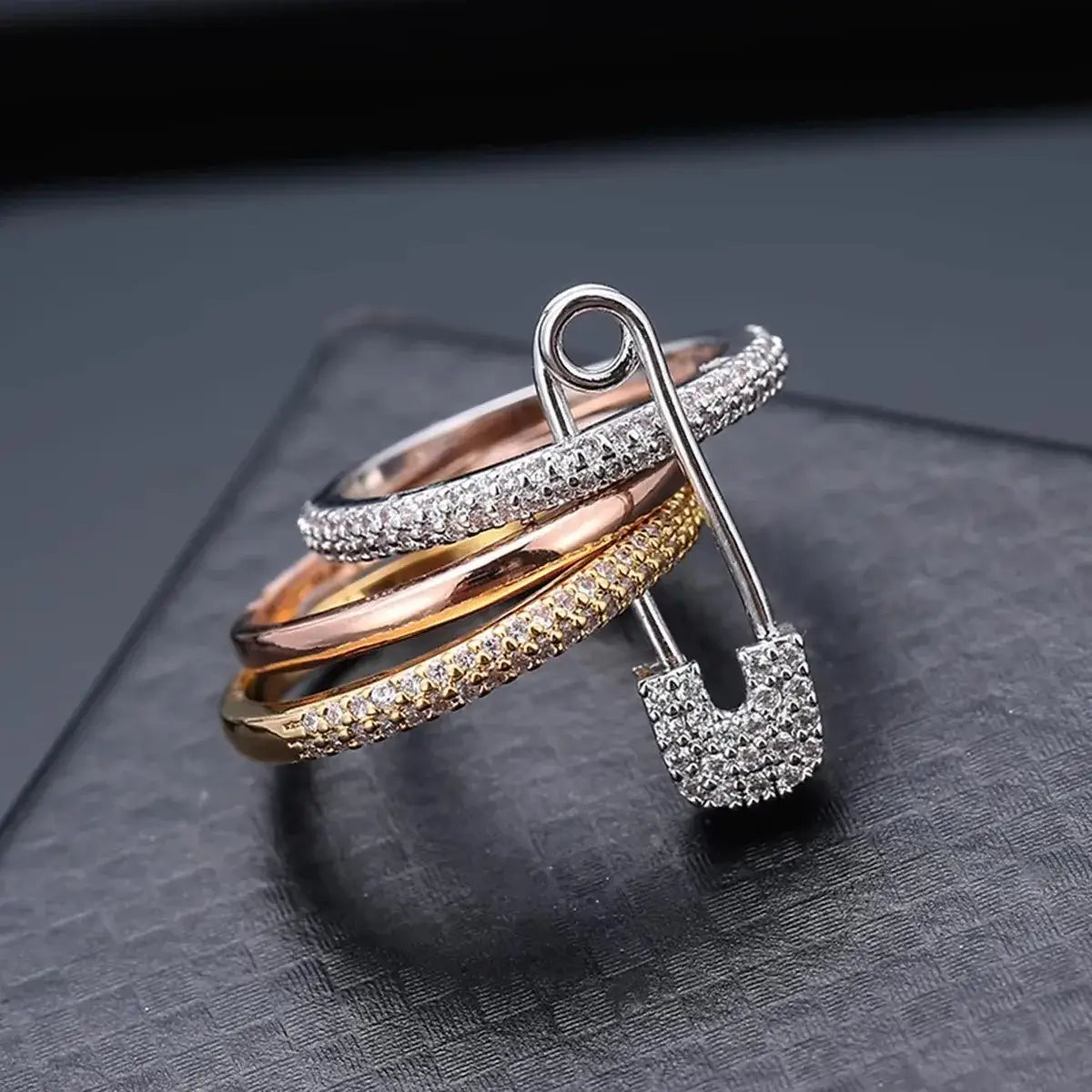 Detailed product close-up on a dark textured surface, showing the silver-tone safety pin, pavé heart charm, and three-tone stacked bands (silver/rose/gold tones) — Tri-Color Safety Pin Ring | OriaJewel