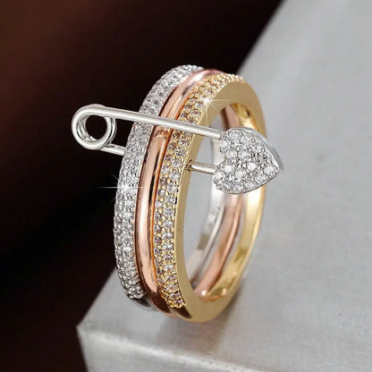 Macro shot on a neutral pedestal capturing the pavé-lined bands, tri-color shine, and the safety pin detail with a heart-shaped pavé charm at the front — Tri-Color Safety Pin Ring | OriaJewel
