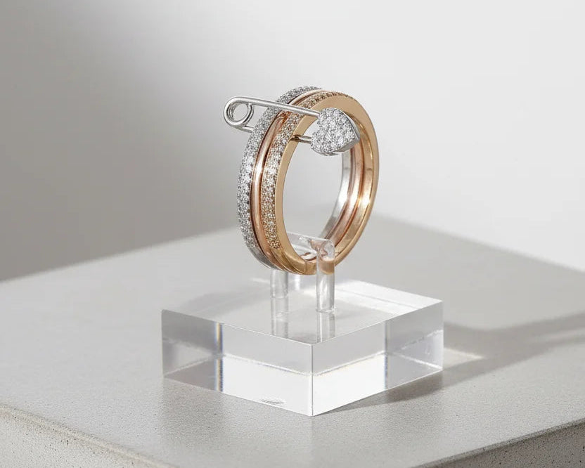 Minimal studio photo with the ring displayed on a clear acrylic stand over a concrete block, emphasizing the tri-color stacked design and safety pin heart charm — Tri-Color Safety Pin Ring | OriaJewel