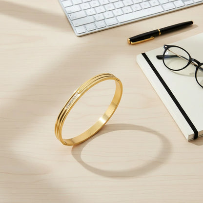 Overhead desk flat lay with a keyboard, notebook, pen, glasses, and the Triple Line Gold Hinged Bangle standing upright, casting a circular shadow on a light wood surface | Oria Jewel