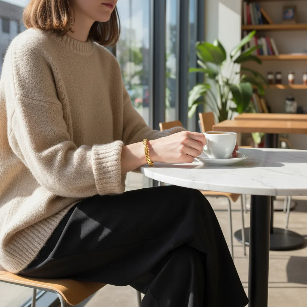 Close-up of a woman sitting at a café table in a soft beige sweater, hands wrapped around a cup while a Twisted Gold Cuff Bracelet shines on her wrist, creating a relaxed everyday coffee-shop look | Oria Jewel