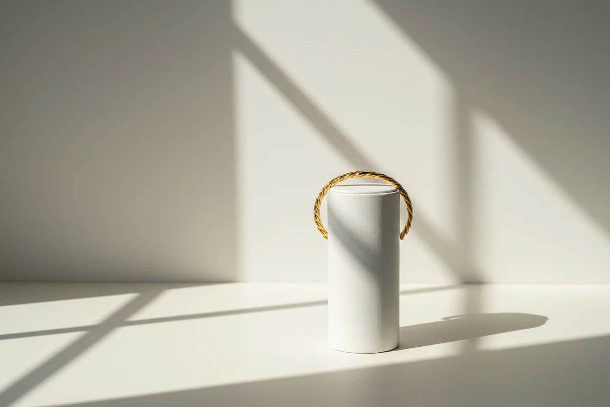 Minimalist hero image of a Twisted Gold Cuff Bracelet draped over a tall white cylindrical stand, captured in strong natural sunlight with dramatic shadows emphasizing its sculpted twist texture | Oria Jewel