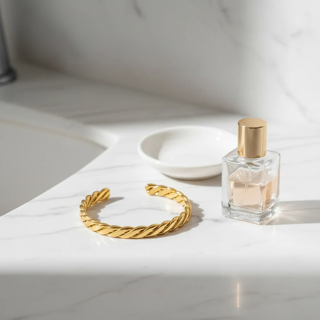 Bathroom vanity scene with a Twisted Gold Cuff Bracelet resting on a white marble countertop next to a glass perfume bottle and ceramic dish, showcasing its elegant twist design in a bright, clean setting | Oria Jewel