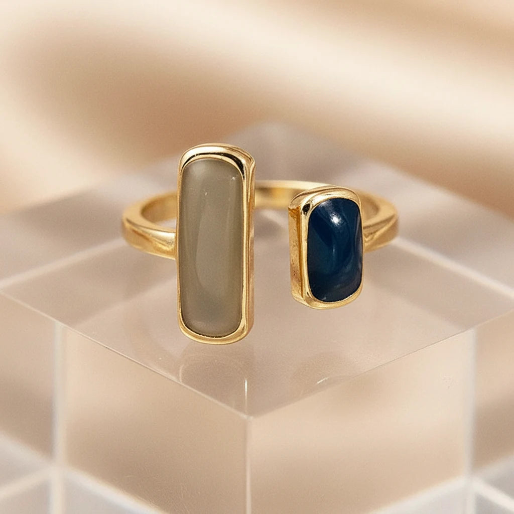 Two Stone Open Ring macro close-up on a clear acrylic block, warm glowing background, gold-tone open band with two rectangular stones (ivory and deep blue) | OriaJewel