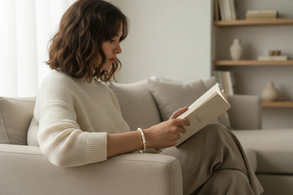 Woman reading on a beige sofa, wearing a delicate White Bamboo bangle Bracelet that adds a soft gold accent to her cozy neutral outfit | Oria Jewel