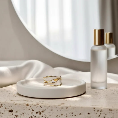 White Enamel Open Wave Ring styled on a minimal vanity scene with a round mirror and frosted bottles, featuring the gold wave detailing over white enamel on a ceramic dish | OriaJewel