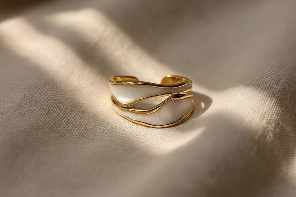 White Enamel Open Wave Ring close-up on beige fabric with soft light patterns, showing the curved gold wave lines and white enamel panels with an open adjustable fit | OriaJewel