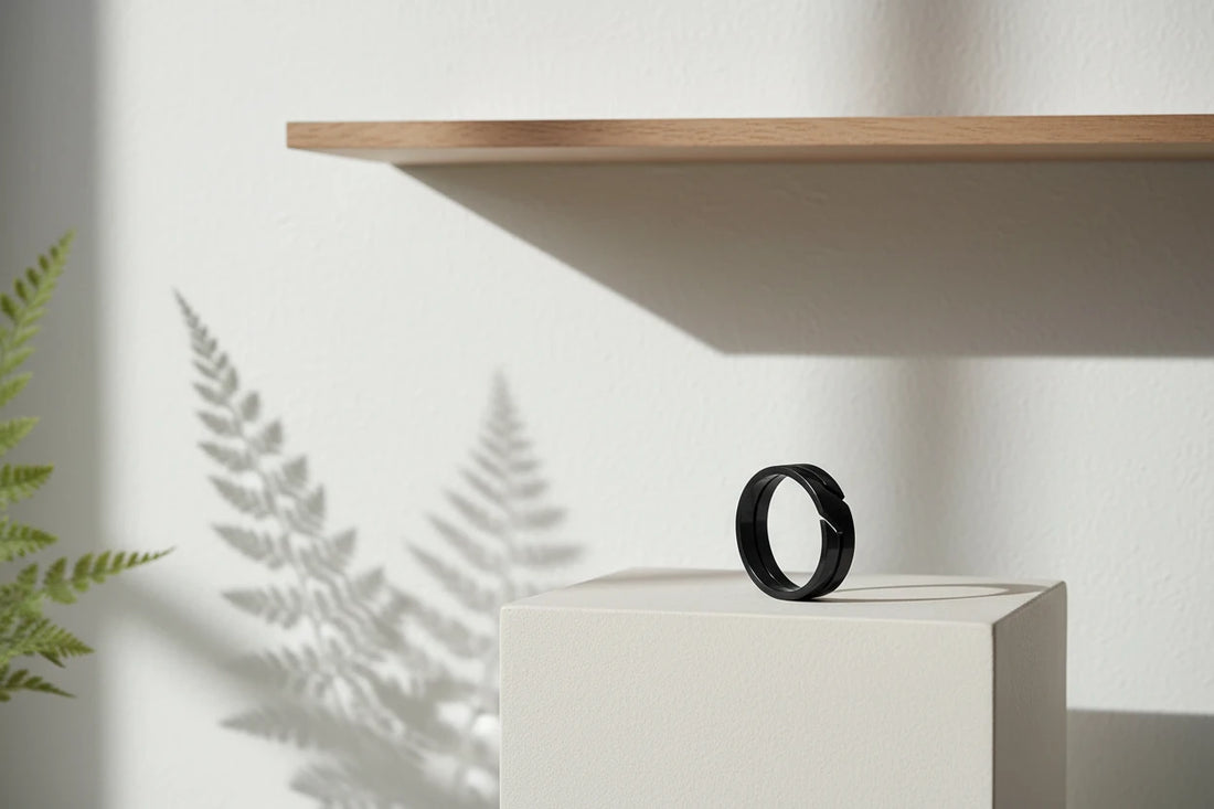Wide Black Band Ring displayed upright on a clean white pedestal under a floating shelf, with soft plant-shadow patterns on the wall to highlight the modern black band silhouette | OriaJewel