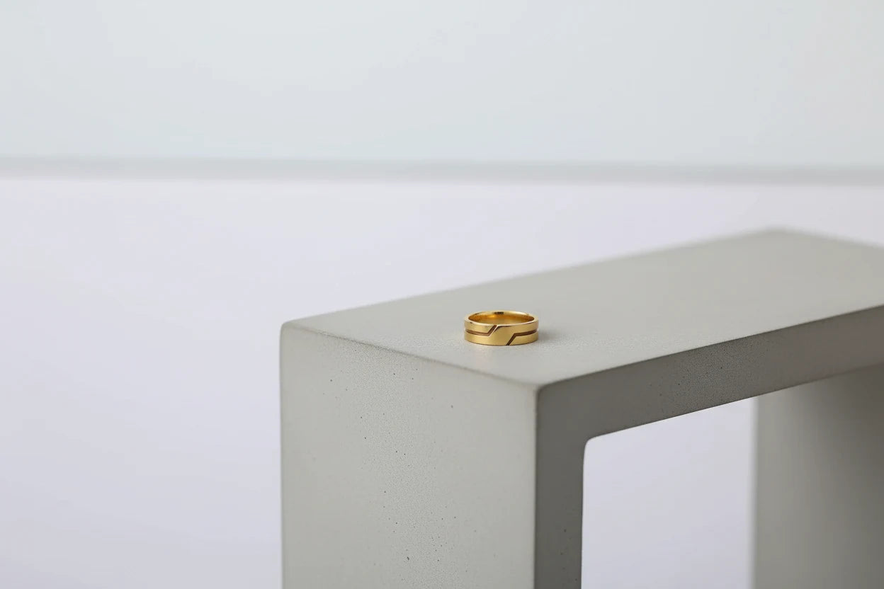 Wide Gold Band Ring placed on the edge of a modern concrete block against a clean neutral background, emphasizing the smooth wide band shape and reflective gold finish | OriaJewel