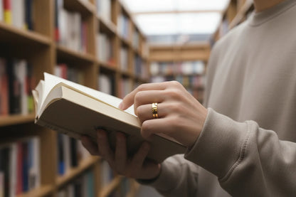 Wide Gold Band Ring worn while browsing a book in a library aisle, close-up on the hand and pages with shelves softly blurred, showing the modern wide band design | OriaJewel
