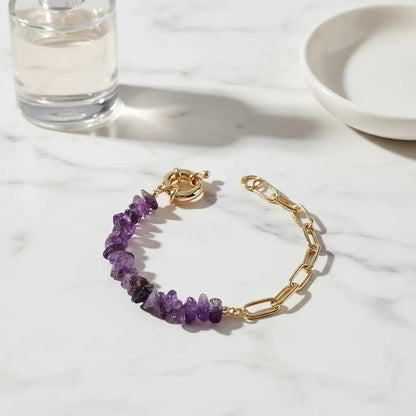 Close-up of a gold paperclip chain and purple gemstone chip amethyst bead bracelet resting on a white marble surface beside a clear perfume bottle and ceramic dish, softly lit to highlight the textured stones and polished metal | Oria Jewel