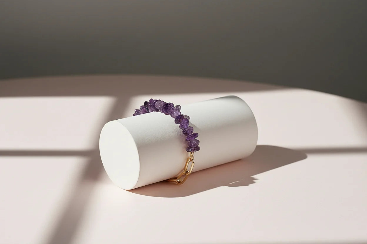 Side view of a purple amethyst bead bracelet draped over a matte white cylinder, combining irregular gemstone chips with a sleek gold paperclip chain on a soft pink surface under dramatic window light and shadow | Oria Jewel