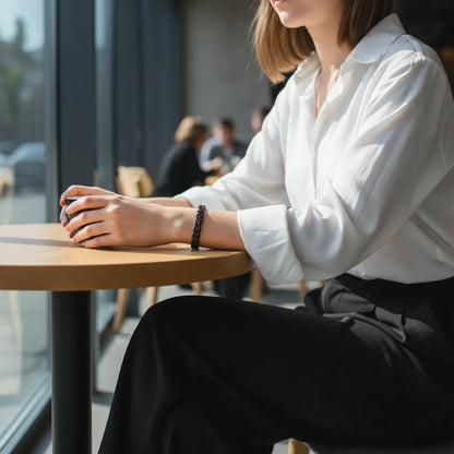 Woman sitting at a café table in soft daylight, wearing a minimalist black chain bracelet on her wrist styled with a white blouse and black trousers, holding a cup with both hands, focus on the sleek black chain bracelet | Oria Jewel