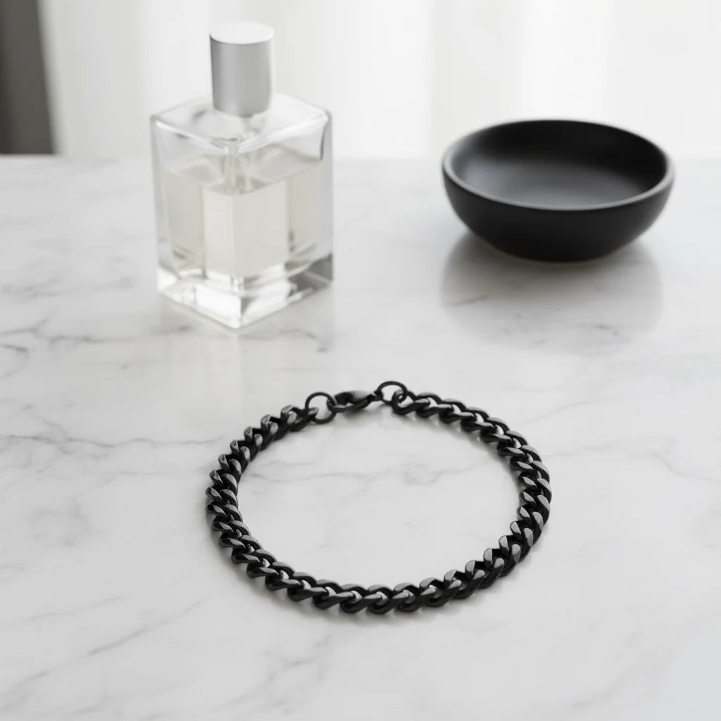 Minimal flat lay of a black chain bracelet on a white marble surface, styled with a clear perfume bottle and a black dish, bright natural light reflecting off the polished black links of the chain bracelet | Oria Jewel