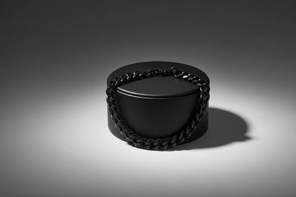 Dramatic studio shot of a black chain bracelet draped over a matte black round jewelry box, spotlight creating strong shadows and reflections that accentuate the bold, masculine look of the black chain bracelet | Oria Jewel