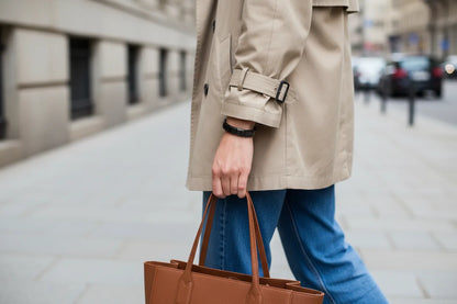 Woman walking in the city wearing a beige trench coat and jeans, holding a brown leather tote bag with a sleek bracelet from our black stainless steel stretch bracelets collection on her wrist, styled for everyday urban chic | Oria Jewel