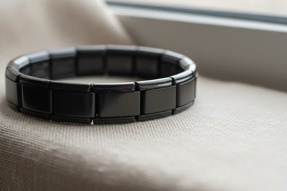 Macro view of a bracelet from the black stainless steel stretch bracelets range resting on soft beige fabric near a window, showcasing the curved rectangular links and modern, high-shine surface in natural daylight | Oria Jewel