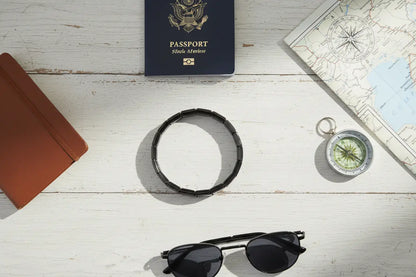 Flat-lay travel scene featuring a bracelet from the black stainless steel stretch bracelets collection arranged with a passport, sunglasses, notebook, compass, and map on a white wooden surface, styled as a versatile accessory for adventures | Oria Jewel