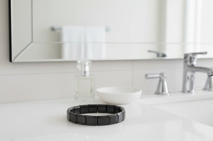 Bathroom counter vignette with a bracelet from the black stainless steel stretch bracelets line placed beside a glass perfume bottle, white dish, and chrome faucet, highlighting its contemporary design in a clean, minimal setting | Oria Jewel