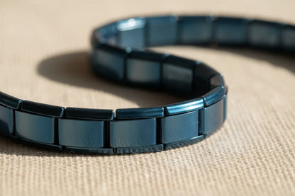 Close-up view of a polished blue stainless steel stretch  bracelets showing individual rectangular links curving on a beige textured fabric surface, with natural sunlight highlighting the smooth metallic shine and flexible design | Oria Jewel