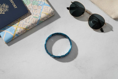 Travel-themed flat lay with a blue stainless steel stretch bracelets placed on a light stone surface beside a folded city map, passport, and round sunglasses, suggesting a stylish accessory for on-the-go adventures | Oria Jewel