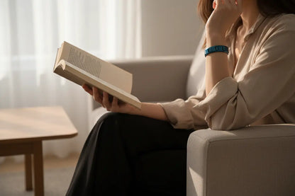 Cozy indoor moment of a woman reading on a sofa near soft curtains, wearing a chic blue stainless steel stretch bracelets that contrasts gently with her neutral blouse and adds a modern accent to her relaxed look | Oria Jewel