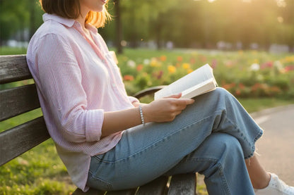 Woman sitting on a wooden park bench reading a book at golden hour, wearing a delicate cz tennis bracelet that sparkles softly against her wrist in the warm sunset light | Oria Jewel