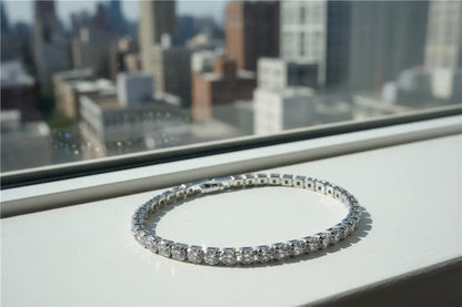 Silver cz tennis bracelet resting on a bright windowsill with a blurred city skyline in the background, each cubic zirconia stone catching the natural daylight and reflecting subtle rainbow flashes | Oria Jewel