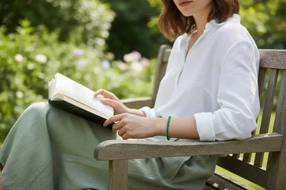 Woman relaxing on a wooden garden bench reading a book, wearing a vivid green cz tennis bracelet that contrasts against her crisp white blouse and soft green skirt in a lush outdoor setting | Oria Jewel