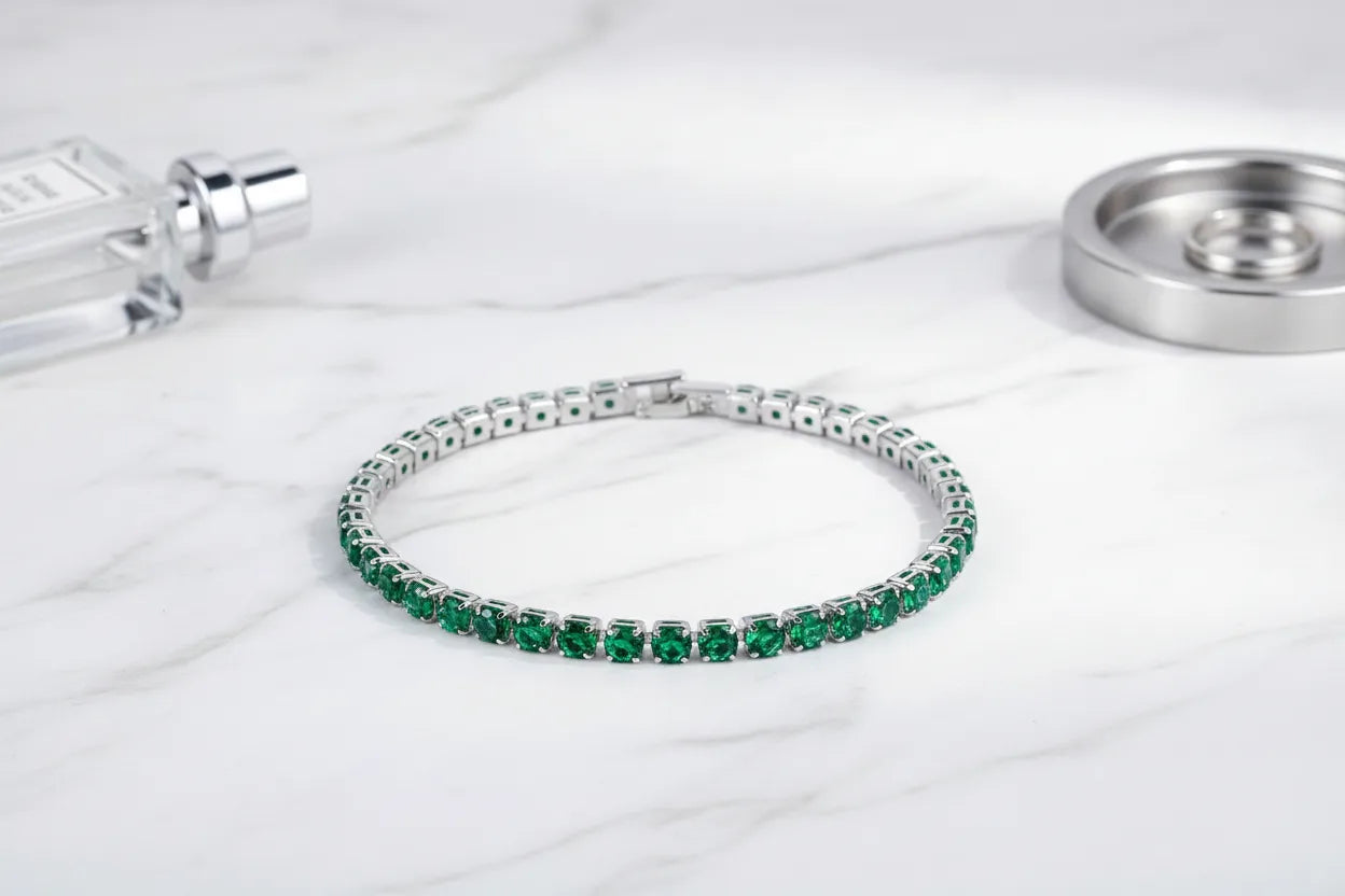 Green cz tennis bracelet displayed in a graceful circle on a white marble surface, styled with a sleek perfume bottle and metal dish, highlighting the rich gemstone color and polished silver setting | Oria Jewel