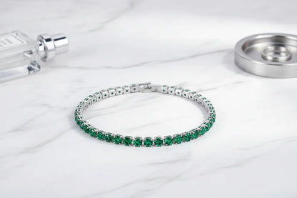 Green cz tennis bracelet displayed in a graceful circle on a white marble surface, styled with a sleek perfume bottle and metal dish, highlighting the rich gemstone color and polished silver setting | Oria Jewel