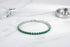 Green cz tennis bracelet displayed in a graceful circle on a white marble surface, styled with a sleek perfume bottle and metal dish, highlighting the rich gemstone color and polished silver setting | Oria Jewel