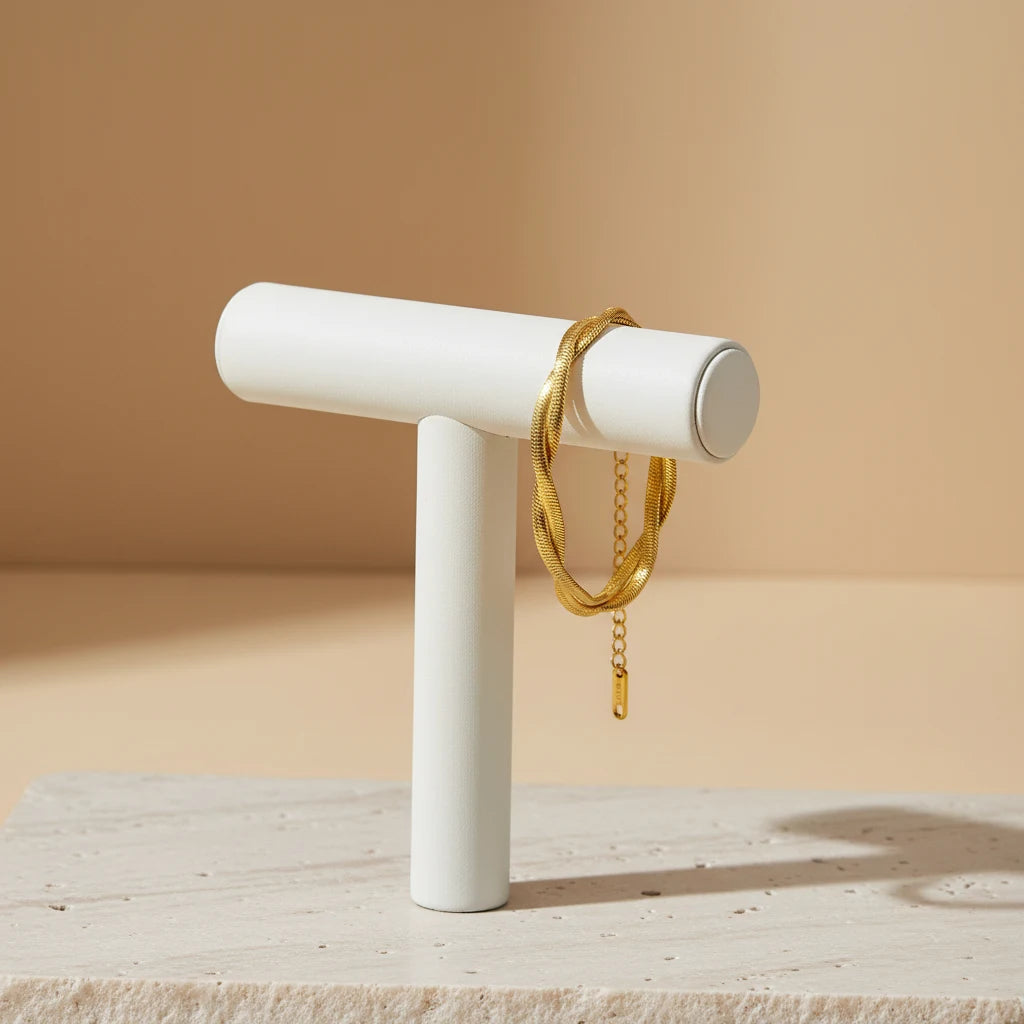 Minimal display of a double chain gold bracelet draped over a modern white T-bar jewelry stand on a textured stone base with warm lighting | Oria Jewel