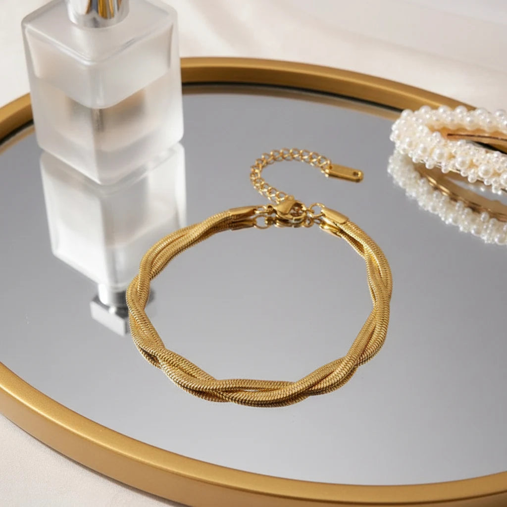 Elegant vanity scene with a double chain gold bracelet resting on a round mirrored tray next to a frosted perfume bottle and pearl accessory | Oria Jewel