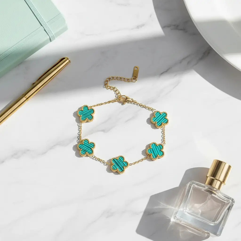 Flat lay of a four leaf clover charm bracelet with turquoise inlay flowers on a delicate gold chain, styled on a white marble surface beside a mint notebook, gold pen, and clear perfume bottle, focus keyword four leaf clover charm bracelet | Oria Jewel