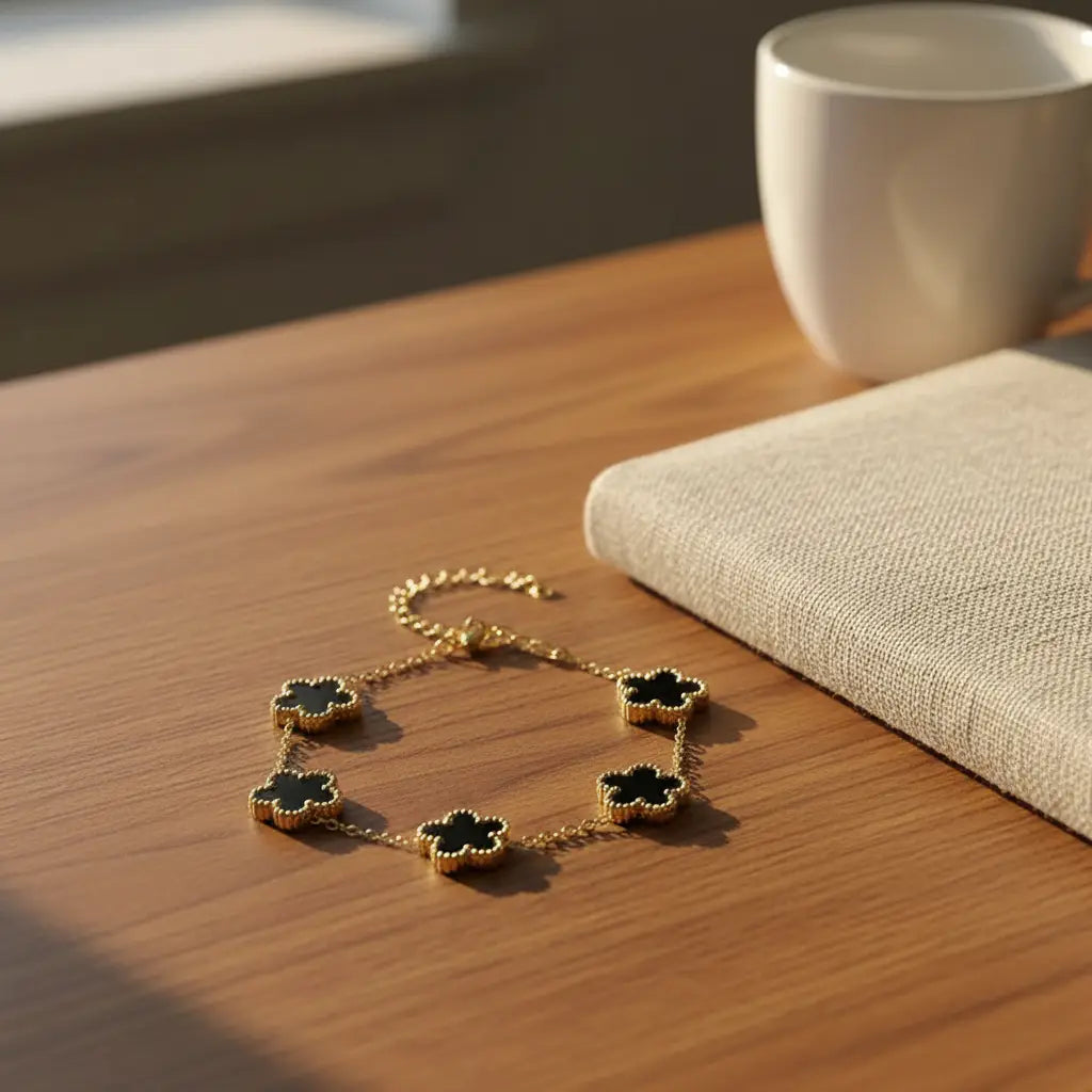 Warm afternoon desk scene with sun streaming through a window, showing a gold four leaf clover charm bracelet with black clover charms lying next to a textured beige notebook and white mug on a wooden surface, focus keyword four leaf clover charm bracelet | Oria Jewel