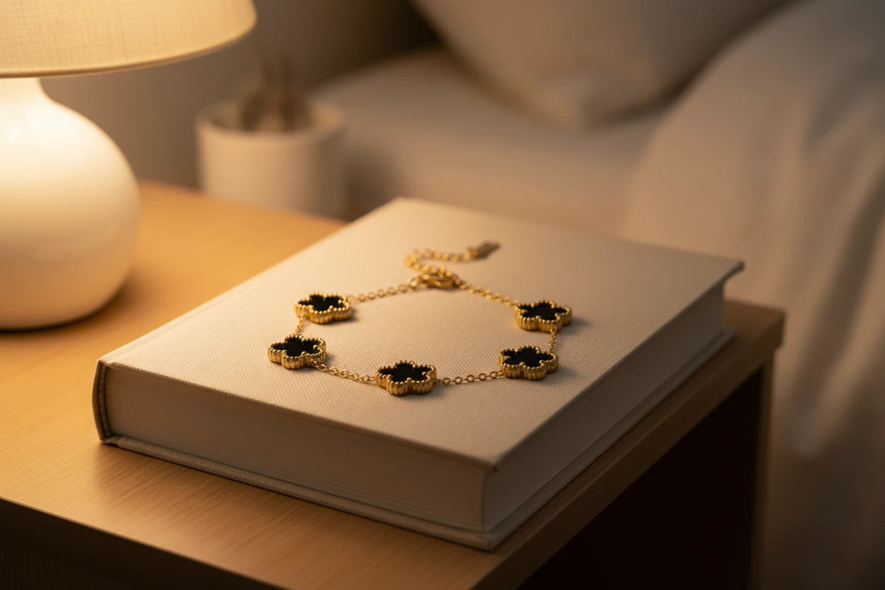 Cozy bedside scene with a gold four leaf clover charm bracelet featuring glossy black clover charms draped over a closed fabric-covered book next to a warm table lamp, focus keyword four leaf clover charm bracelet | Oria Jewel