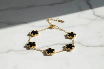 Close-up of a gold four leaf clover charm bracelet with glossy black clover charms arranged in a gentle curve on a white marble background with diagonal sunlight and soft shadows, focus keyword four leaf clover charm bracelet | Oria Jewel