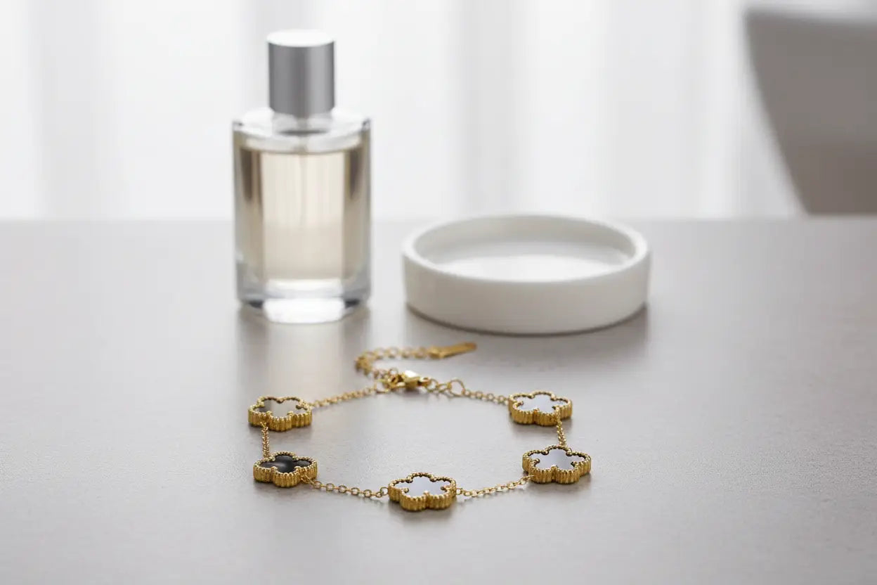 Minimal beauty setup with a gold four leaf clover charm bracelet featuring alternating black and white clover charms on a grey tabletop beside a clear perfume bottle and white trinket dish, focus keyword four leaf clover charm bracelet | Oria Jewel