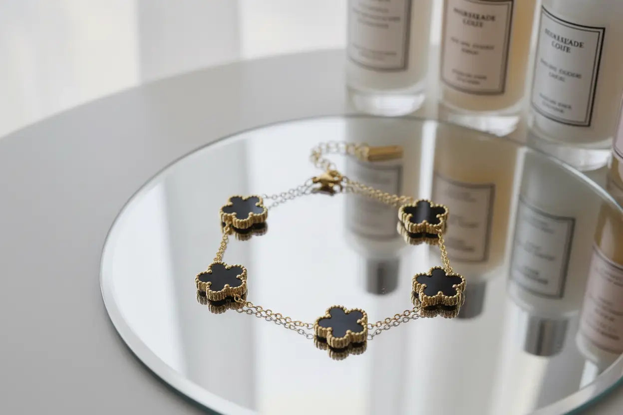 Elegant styling scene with a gold four leaf clover charm bracelet with black enamel flowers displayed on a round mirror, surrounded by tall scented candle bottles reflected in the surface, focus keyword four leaf clover charm bracelet | Oria Jewel