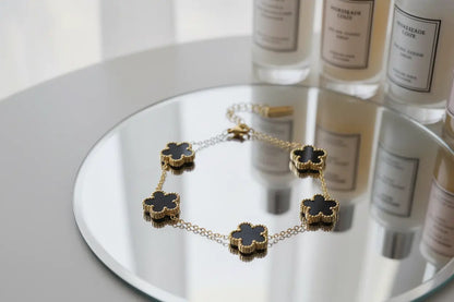 Elegant styling scene with a gold four leaf clover charm bracelet with black enamel flowers displayed on a round mirror, surrounded by tall scented candle bottles reflected in the surface, focus keyword four leaf clover charm bracelet | Oria Jewel