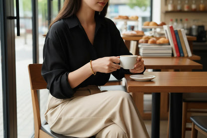 Woman sitting in a cozy café, wearing a black blouse and beige trousers, holding a coffee cup with a sleek gold Bamboo bangle Bracelet on her wrist, casual chic daytime style | Oria Jewel