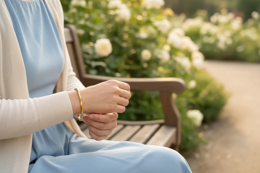 Close-up of a woman on a park bench in a light blue dress and cream cardigan, gently adjusting her gold Bamboo bangle Bracelet surrounded by soft-focus blooming flowers | Oria Jewel