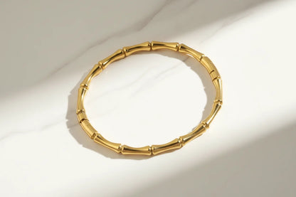 Solo product shot of a shiny gold Bamboo bangle Bracelet laid flat on white marble, highlighted by natural sunlight and soft shadows to show its curved bamboo links | Oria Jewel