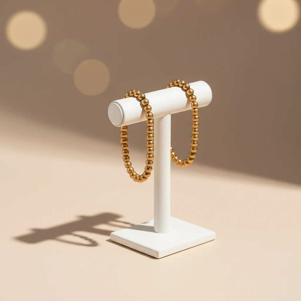 Minimalist display of a gold beaded stretch bracelet draped over a modern white jewelry stand against a soft beige background with warm bokeh light, highlighting its polished spheres | Oria Jewel