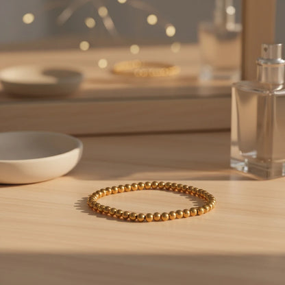Gold beaded stretch bracelet resting on a light wooden vanity beside a mirror, perfume bottle, and soft fairy lights, styled as an elegant daily accessory | Oria Jewel