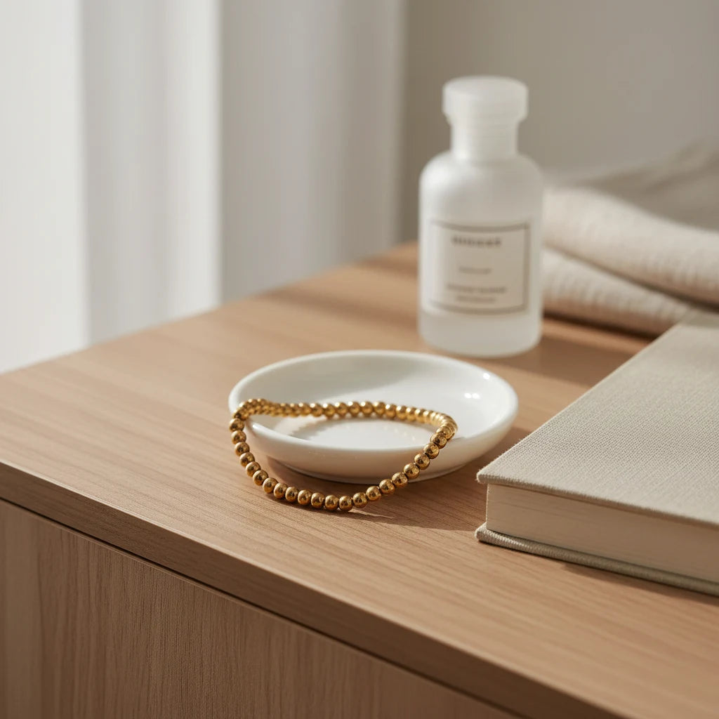 Gold beaded stretch bracelet placed in a small white trinket dish on a wooden bedside table next to a fabric-covered book and skincare bottle, suggesting a calm morning routine | Oria Jewel