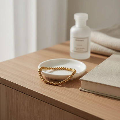 Gold beaded stretch bracelet placed in a small white trinket dish on a wooden bedside table next to a fabric-covered book and skincare bottle, suggesting a calm morning routine | Oria Jewel